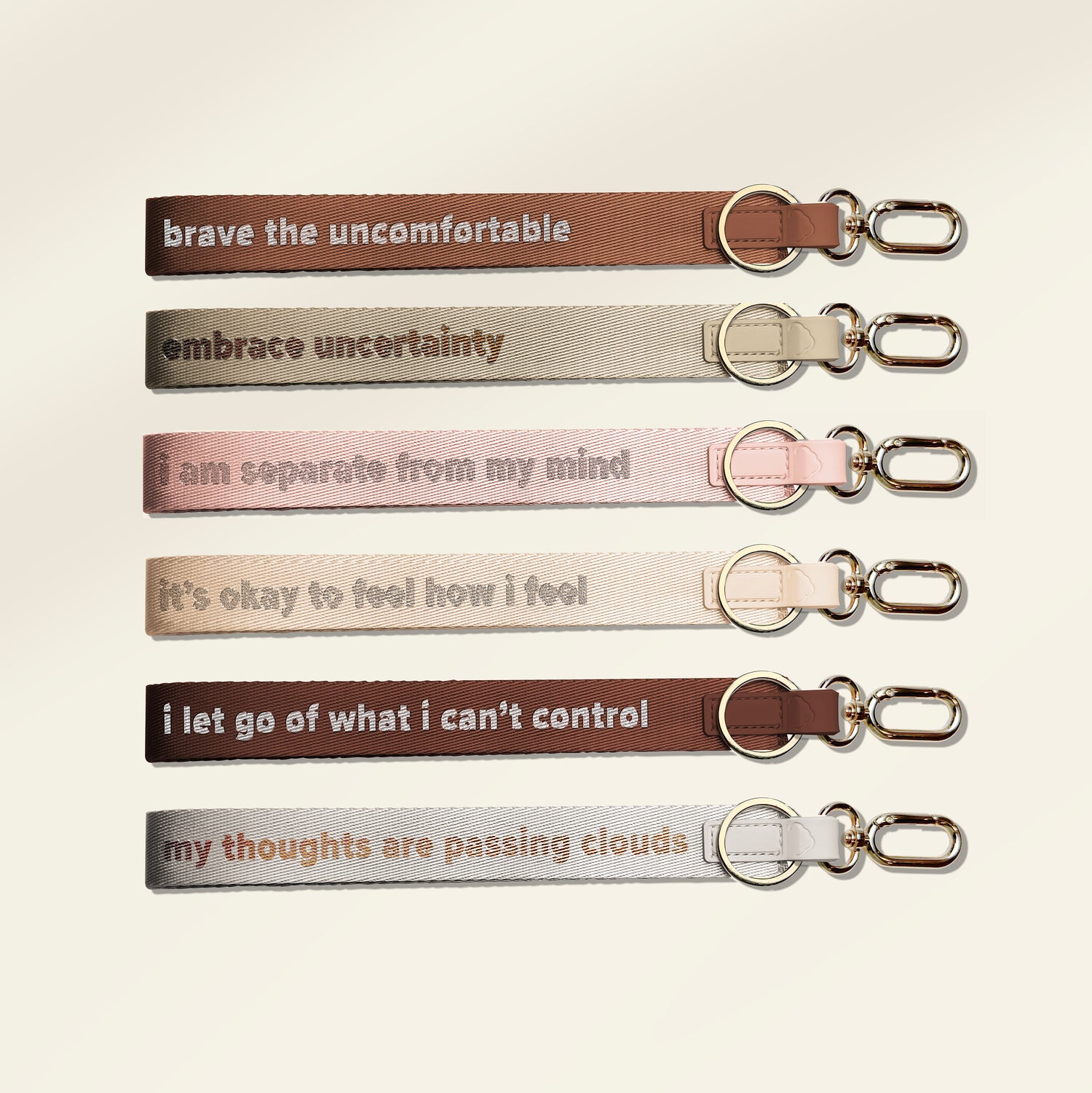 Six fabric keychains with silver metal keyrings and clips, arranged vertically on a light cream background. Each keychain has a different color and a supportive quote printed in lowercase serif text: 'i let go of what i can't control', 'brave the uncomfortable', 'i am separate from my mind', 'it's okay to feel how i feel', 'embrace uncertainty', and 'my thoughts are passing clouds'.