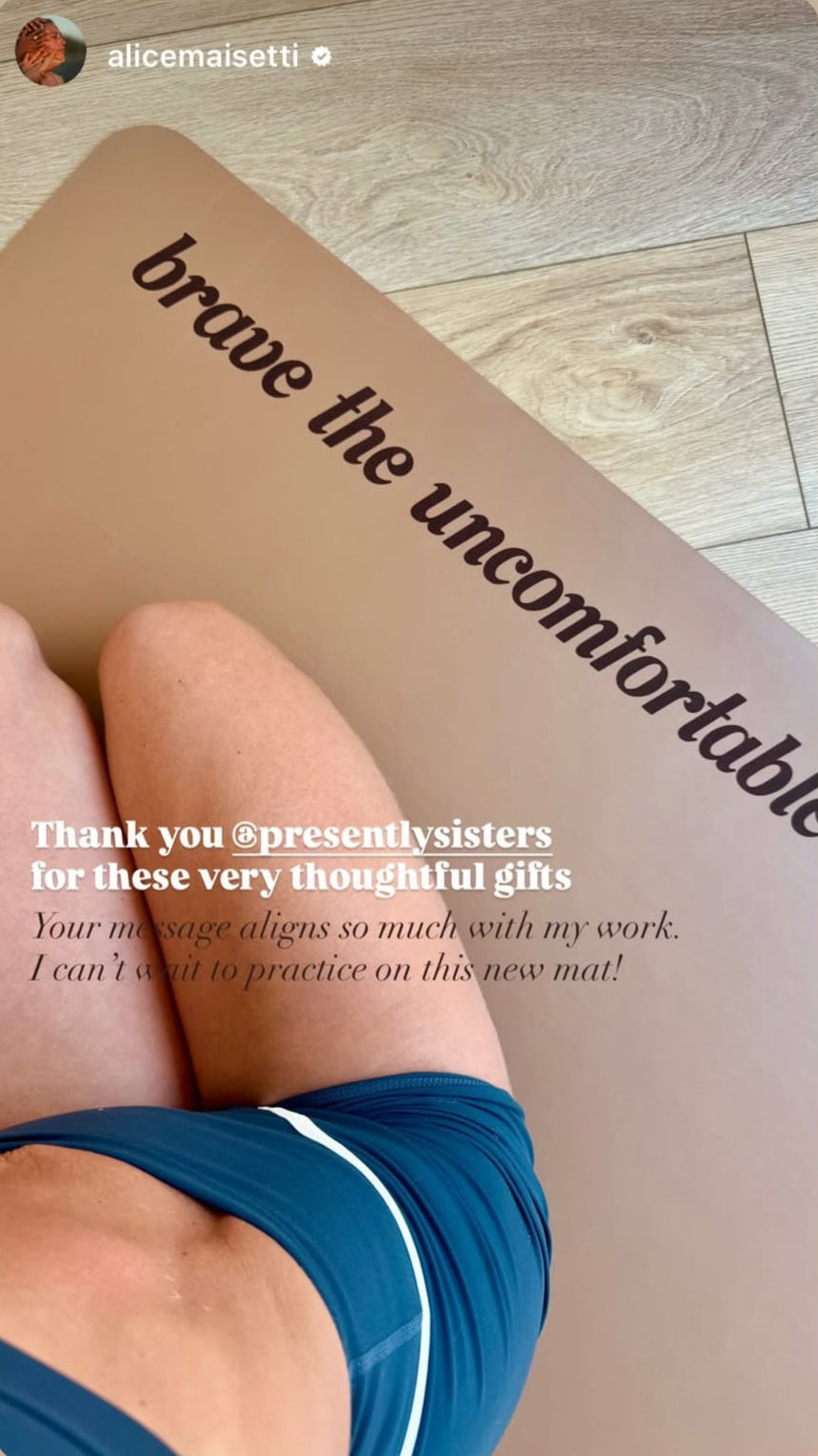 A close-up photo of a person lying on a tan yoga mat with the phrase 'brave the uncomfortable' printed in black script. The person is wearing blue athletic bottoms, and the scene is photographed on a light wood floor.