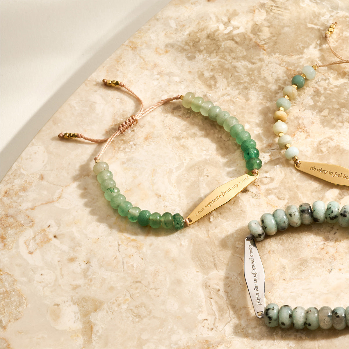 Close-up of an Aventurine gemstone bracelet next to other beaded bracelets from Presently's collection, showcasing the engraved phrase 