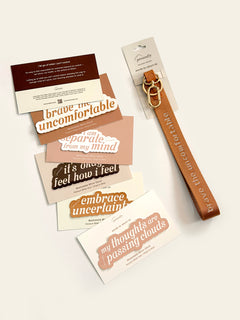 A flat-lay product photo of a brown nylon keychain with gold hardware and matching printed stickers/cards in earthy tones, arranged on a light beige background. The keychain has the words 'brave the uncomfortable' printed on it, and the cards feature phrases like 'brave the uncomfortable', 'I am separate from my mind', and 'my thoughts are passing clouds'.