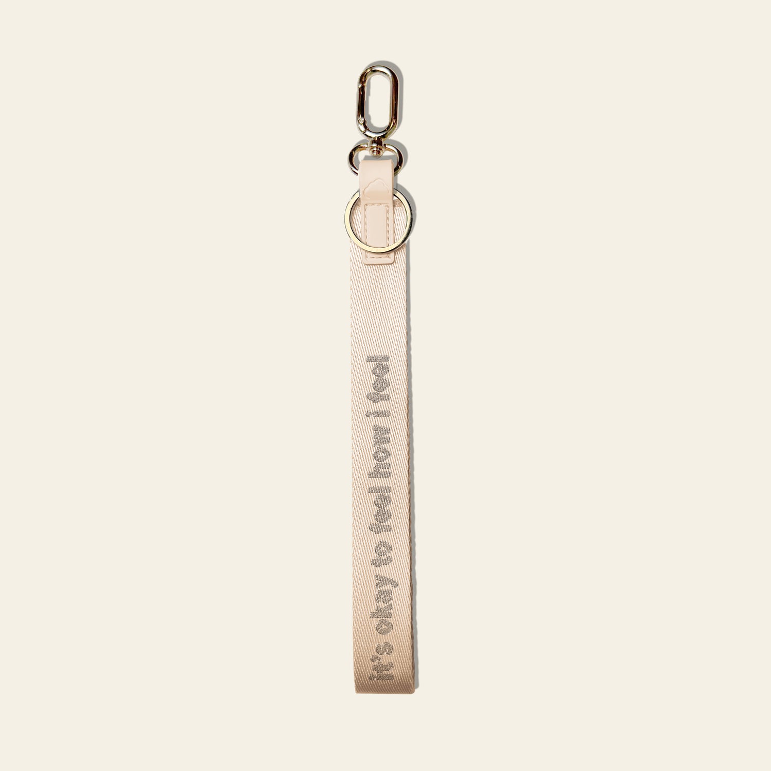A light beige fabric keychain strap with the text 'it's okay to feel how i feel' printed vertically in a darker beige font, attached to a silver metal lobster clasp and split ring, on a solid off-white background.