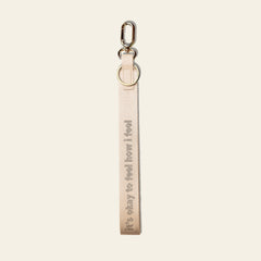 A light beige fabric keychain strap with the text 'it's okay to feel how i feel' printed vertically in a darker beige font, attached to a silver metal lobster clasp and split ring, on a solid off-white background.