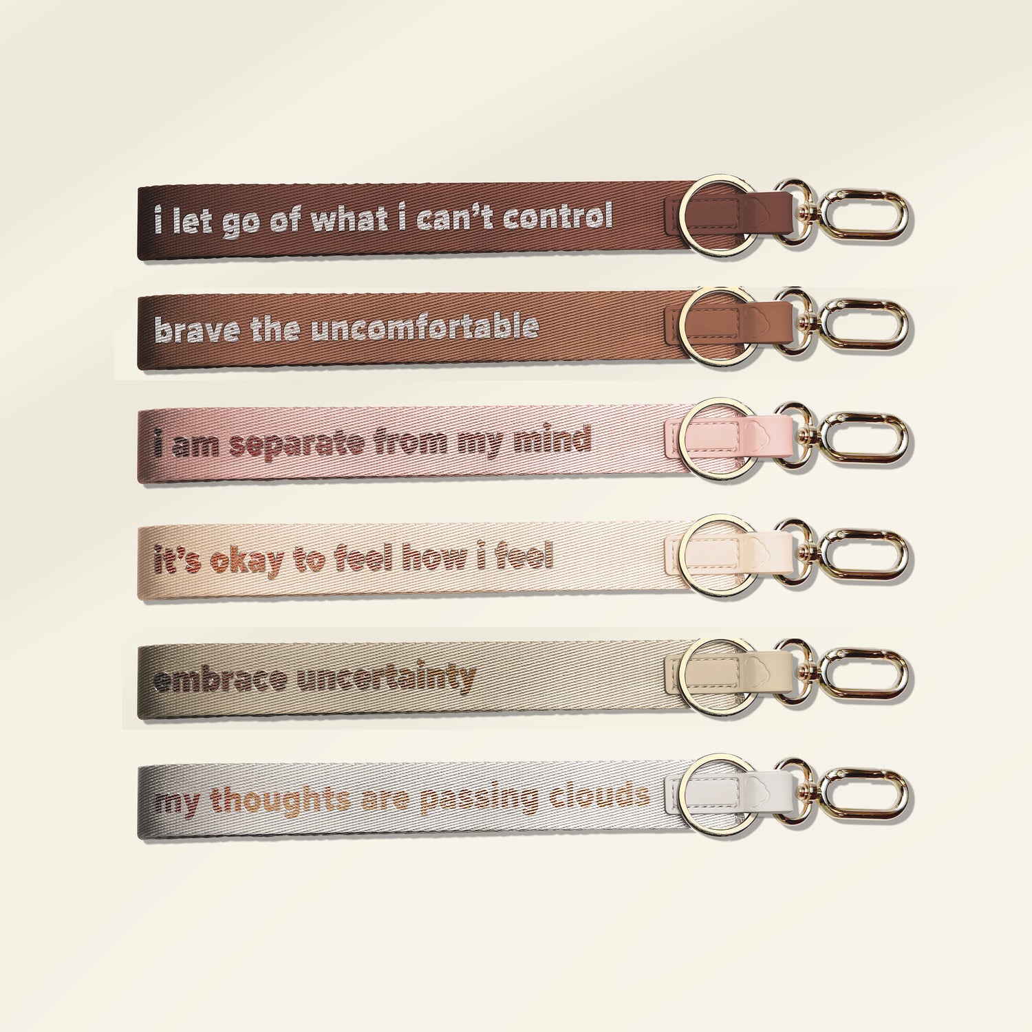 Six fabric keychains with silver metal keyrings and clips, arranged vertically on a light cream background. Each keychain has a different color and a supportive quote printed in lowercase serif text: 'i let go of what i can't control', 'brave the uncomfortable', 'i am separate from my mind', 'it's okay to feel how i feel', 'embrace uncertainty', and 'my thoughts are passing clouds'.