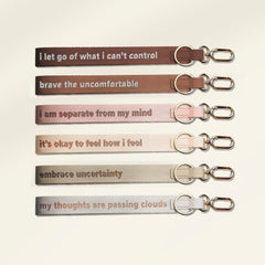 Six fabric keychains with silver metal keyrings and clips, arranged vertically on a light cream background. Each keychain has a different color and a supportive quote printed in lowercase serif text: 'i let go of what i can't control', 'brave the uncomfortable', 'i am separate from my mind', 'it's okay to feel how i feel', 'embrace uncertainty', and 'my thoughts are passing clouds'.