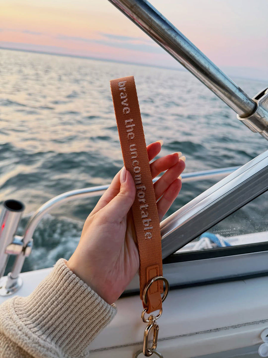 Brown “Brave the Uncomfortable” keychain wristlet held on a boat at sunset